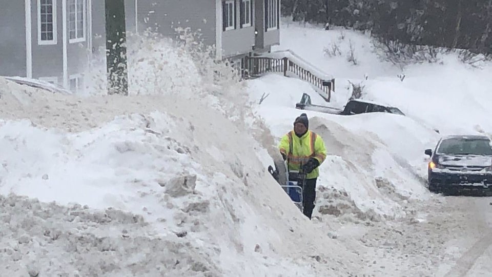 Un homme manie sa souffleuse devant un énorme amoncellement de neige. La neige est plus haute que les voitures garées dans la rue.